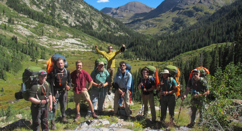 A group of people wearing backpacks pose for a photo in green mountainous valley.